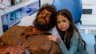 Paulo interpretado por Elam Lima ferido em uma maca ao lado de Anna vivida por Mel Summers em cena da novela A Caverna Encantada