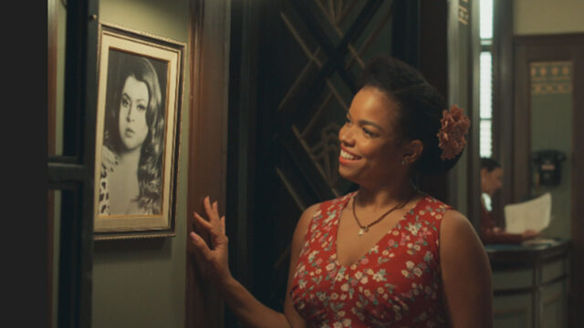 Dita (Jeniffer Nascimento) sorri enquanto observa um retrato em preto e branco de Preta Gil na novela Êta Mundo Melhor!