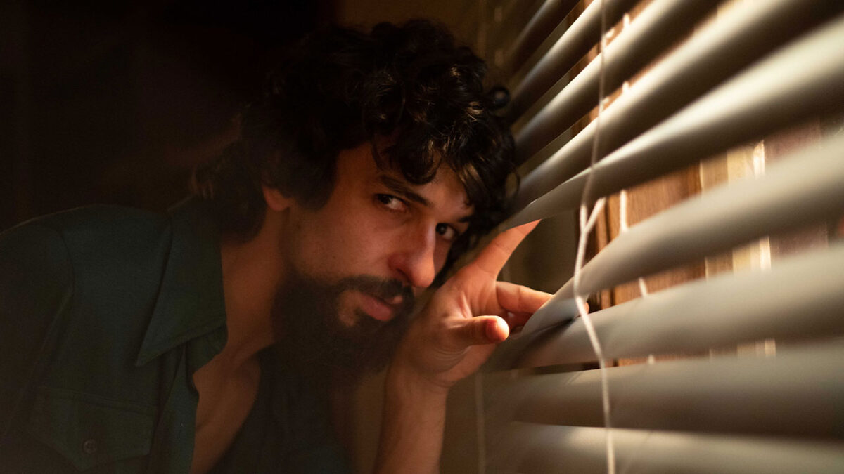 Ravel Andrade caracterizado como Raul Seixas observando pela persiana com expressão séria em cena da cinebiografia do cantor