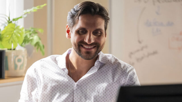 Ricardo Burgos sorrindo enquanto olha para frente usando camisa social branca estampada em ambiente de escritório com quadro branco e planta ao fundo
