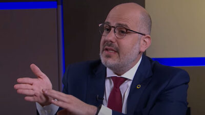 Andre Basbaum fala durante entrevista usando terno azul escuro, camisa branca e gravata vermelha em estúdio de TV