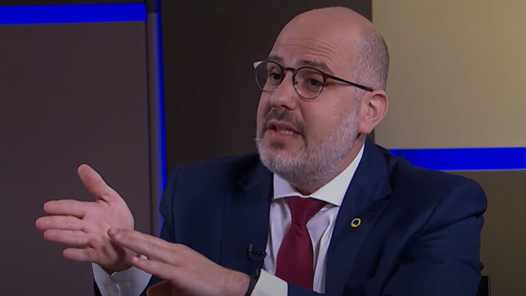Andre Basbaum fala durante entrevista usando terno azul escuro, camisa branca e gravata vermelha em estúdio de TV