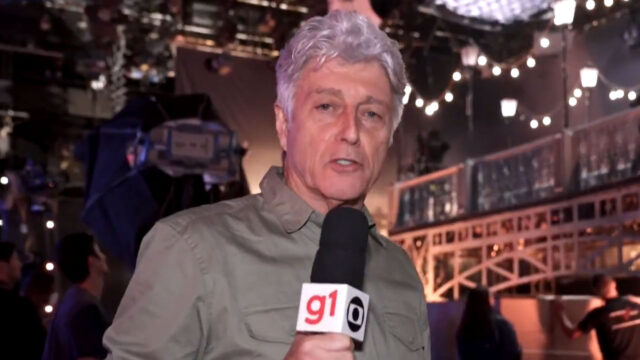 Caco Barcellos segura microfone da Globo em estúdio iluminado com refletores e cenário decorado ao fundo