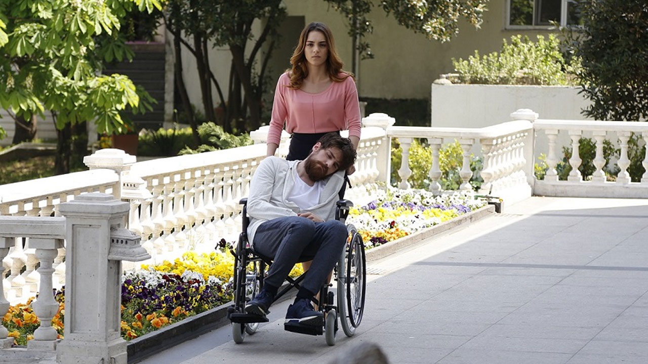 Cemre (Sera Kutlubey) empurra Nedim (Berker Guven) em cadeira de rodas em jardim na novela Cruel Istambul