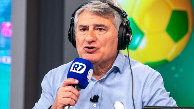 Cléber Machado com fones de ouvido narra ao microfone do R7 em estúdio esportivo da Record com imagem de bola de futebol ao fundo