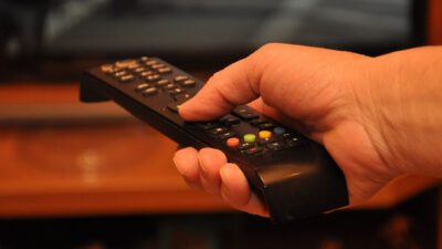 Mão segura controle remoto preto apontado para televisão ligada em ambiente doméstico