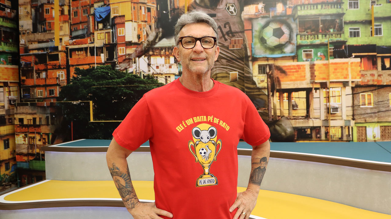 Craque Neto com óculos e camiseta vermelha com estampa de troféu no estúdio do programa Os Donos da Bola