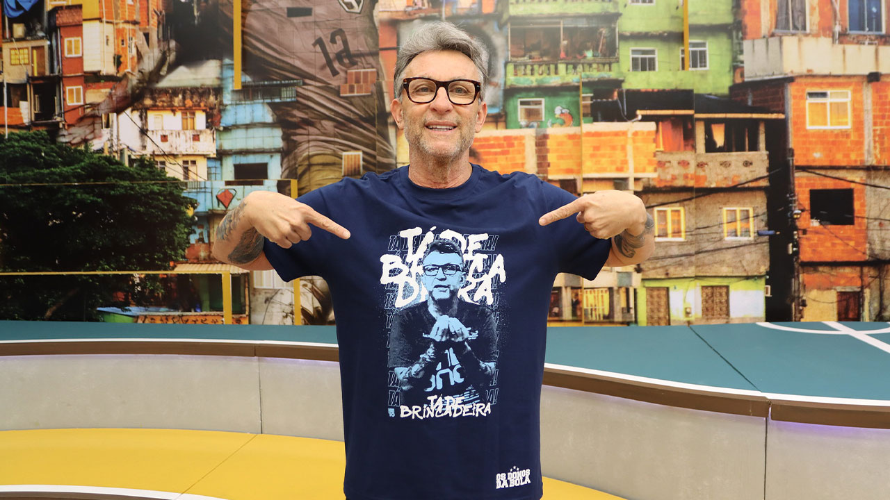 Craque Neto de óculos aponta para camiseta azul com estampa de sua imagem no estúdio do programa Os Donos da Bola