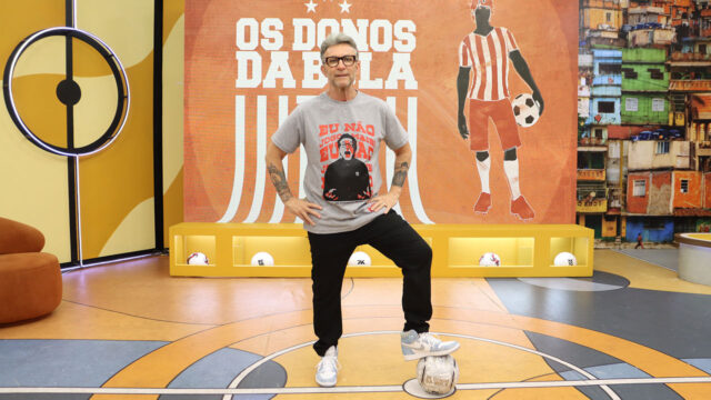 Craque Neto com óculos e camiseta cinza pisa em bola no estúdio do programa Os Donos da Bola
