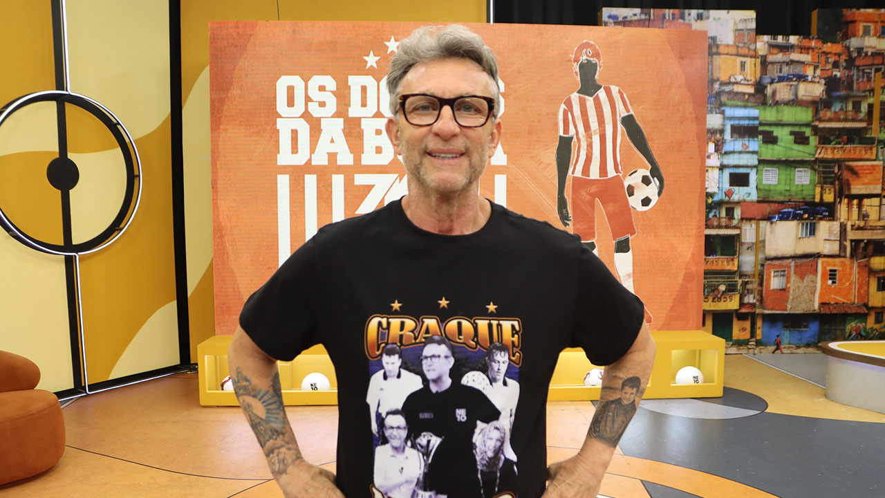 Craque Neto com óculos e camiseta preta estampada com fotos no estúdio do programa Os Donos da Bola