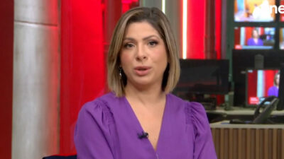 Daniela Lima com blusa roxa apresenta o Conexão GloboNews em estúdio com telas e luzes vermelhas ao fundo