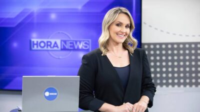 Apresentadora Daysa Belini sorrindo, de blazer preto, em estúdio da Record News com tela azul exibindo o logo Hora News