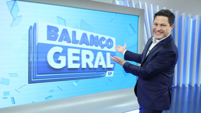 Eleandro Passaia sorri de terno escuro e aponta para o telão com o logotipo do Balanço Geral SP no estúdio