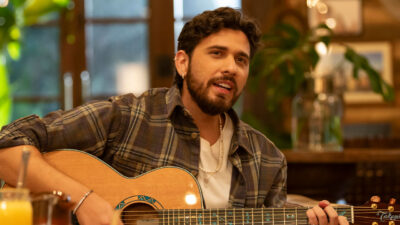 Gustavo Mioto de camisa xadrez e colar de pérolas cantando e tocando violão em performance acústica