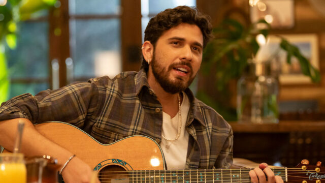 Gustavo Mioto de camisa xadrez e colar de pérolas cantando e tocando violão em performance acústica