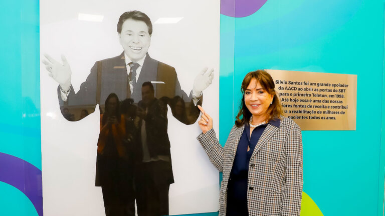 Iris Abravanel sorri ao lado de painel com foto de Silvio Santos durante homenagem ao apresentador na sede da AACD em São Paulo