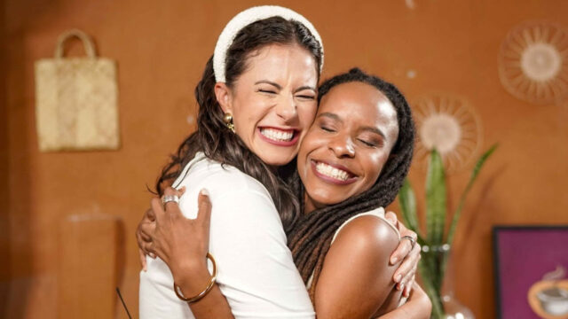 Jéssica Senra e Monique Evelle se abraçam sorrindo com olhos fechados em ambiente interno decorado com plantas e quadros