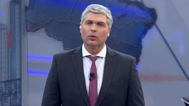 Joel Datena de terno escuro e gravata vermelha apresenta o Brasil Urgente no estúdio da Band com mapa do Brasil ao fundo