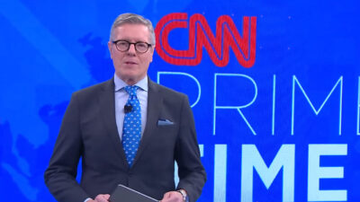 Márcio Gomes de terno escuro e gravata azul apresenta o CNN Prime Time em frente ao telão com o logotipo do telejornal