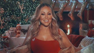 Mariah Carey sorrindo com vestido vermelho em cenário natalino com árvore decorada lareira e meias penduradas ao fundo