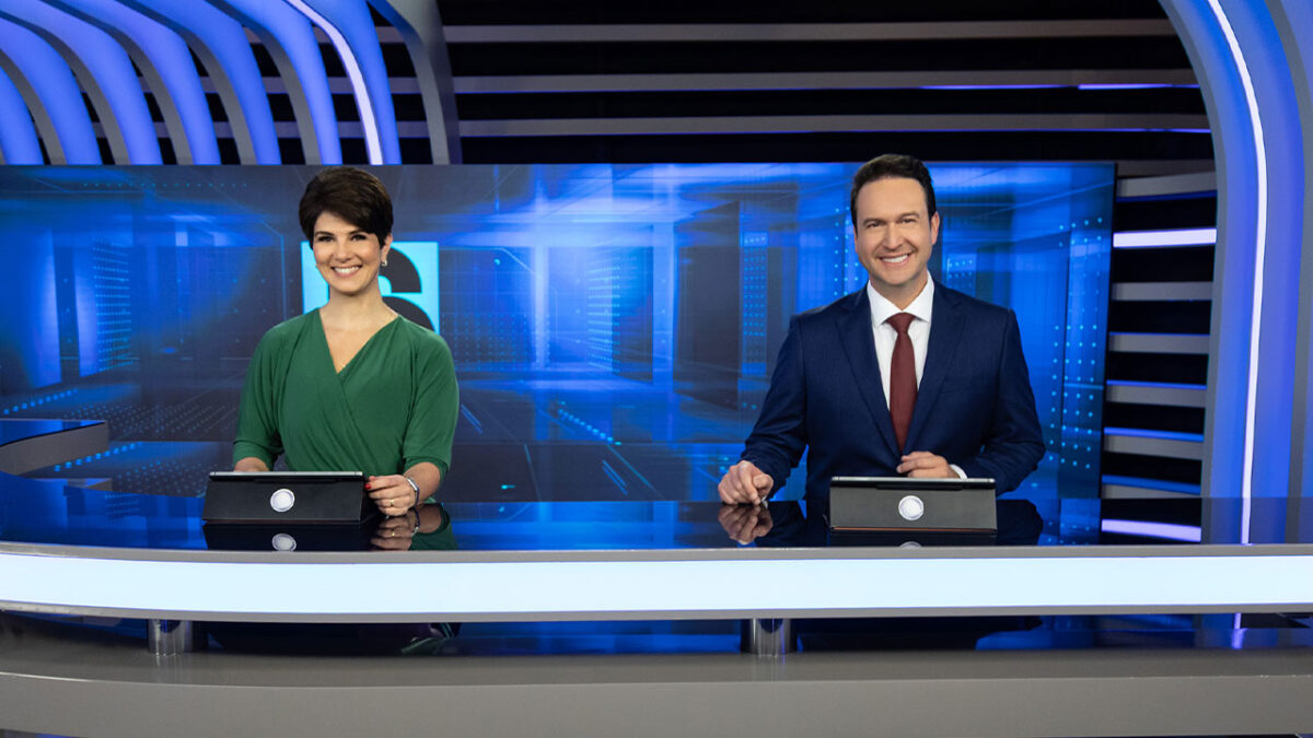 Mariana Godoy de vestido verde e Eduardo Ribeiro de terno azul com gravata vermelha apresentam o Jornal da Record no estúdio