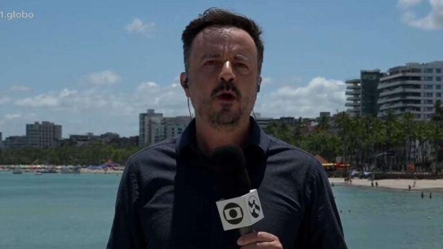 Jornalista Mauro Wedekin segura microfone da TV Globo e faz entrada ao vivo em praia urbana de Maceió com prédios ao fundo