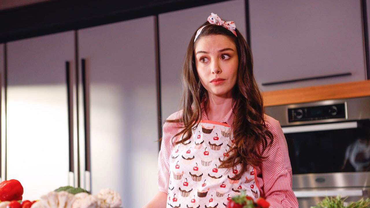 Nazli interpretada por Özge Gürel usa avental estampado e faixa no cabelo em cena na cozinha da novela turca Dolunay