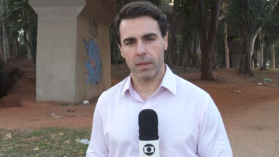 Rafael Colombo segura microfone da TV Globo em reportagem ao ar livre sob viaduto com árvores ao fundo