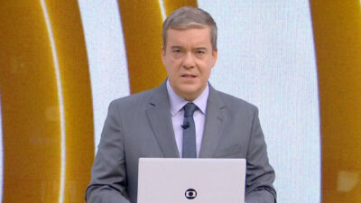 Roberto Kovalick apresenta o telejornal Hora Um da TV Globo usando terno cinza e gravata em frente ao telão do estúdio com logotipo dourado ao fundo