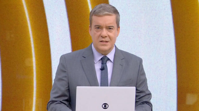 Roberto Kovalick apresenta o telejornal Hora Um da TV Globo usando terno cinza e gravata em frente ao telão do estúdio com logotipo dourado ao fundo