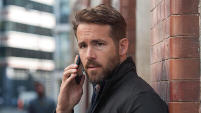 Ryan Reynolds caracterizado como Bill Pope fala ao celular encostado em parede de tijolos em cena do filme Mente Criminosa 2016