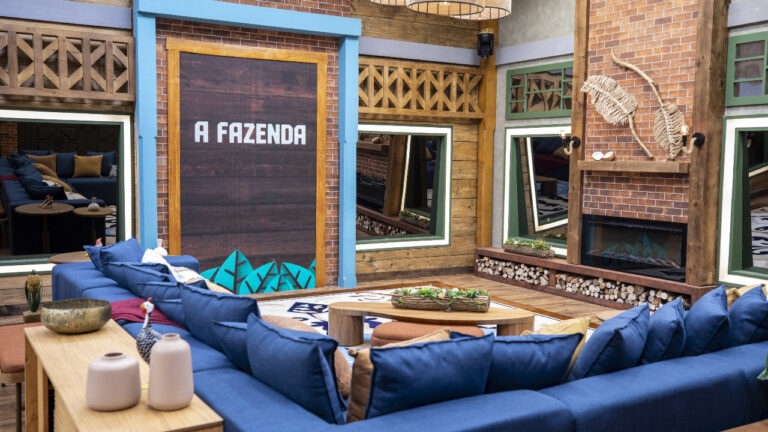 Sala da sede de A Fazenda 17 com sofá azul em formato de U, mesa de centro de madeira e painel de LED com o nome do reality show