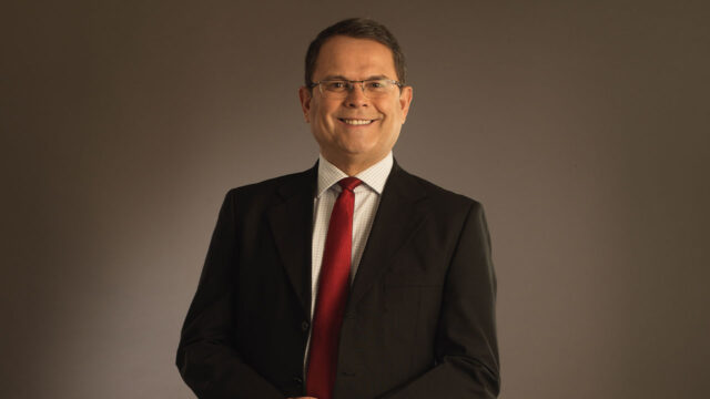 Sidney Rezende sorrindo com terno preto, camisa clara e gravata vermelha em foto com fundo neutro