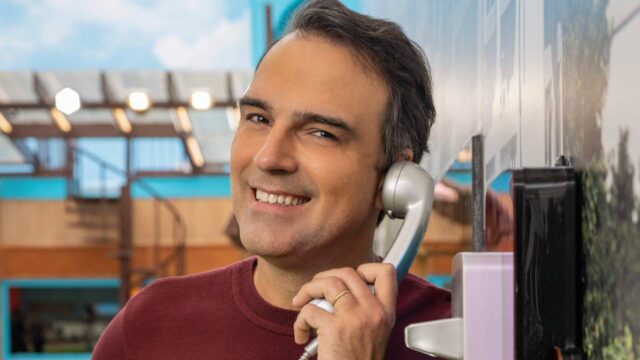 Apresentador Tadeu Schmidt sorrindo, usando suéter vinho e segurando um telefone fixo prateado, em cenário de reality show