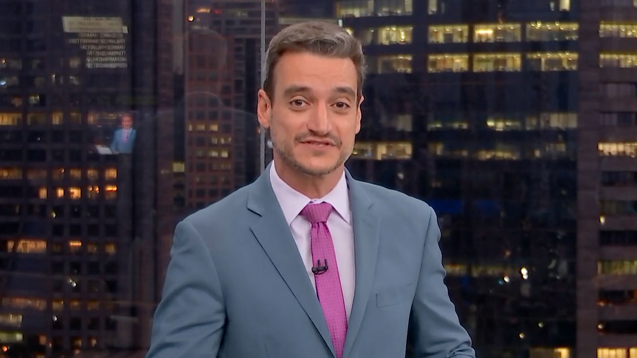 Tiago Scheuer apresenta o telejornal Bom Dia São Paulo usando terno azul claro e gravata rosa com cenário noturno da cidade ao fundo
