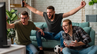 Três homens comemoram assistindo jogo de futebol na TV sentados em sala com sofá verde e bola no chão