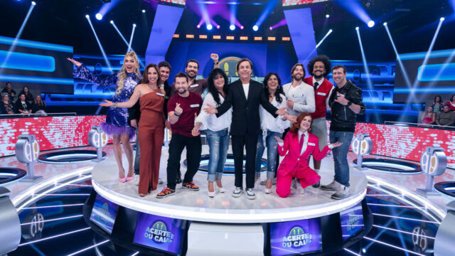 Tom Cavalcante e os participantes do programa Acerte ou Caia! posando sorridentes no centro do palco iluminado, cercados por luzes coloridas e telões com o logotipo da atração