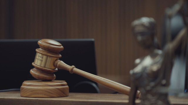 Martelo do juiz em primeiro plano sobre a bancada de um tribunal, com a estátua da Justiça desfocada ao fundo na série Acusado: Fui Eu Que Fiz Isso?