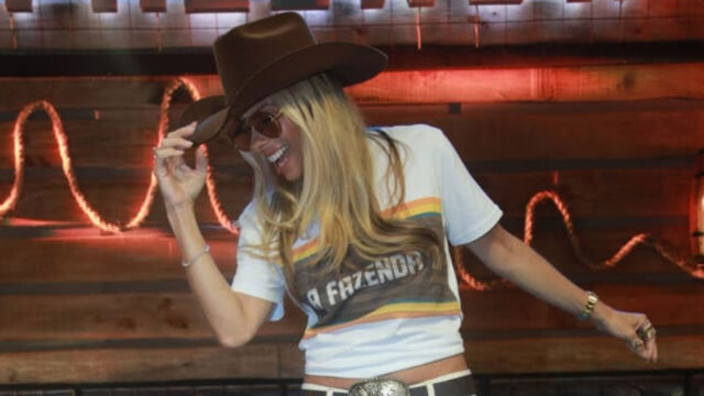 A apresentadora Adriane Galisteu com chapéu de cowboy e camiseta de A Fazenda sorri no cenário rústico do reality show da Record