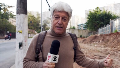 Caco Barcellos segurando microfone com o logotipo do G1 e da TV Globo enquanto faz reportagem de rua com mochila nas costas e área em obras ao fundo em cena do programa Profissão Repórter