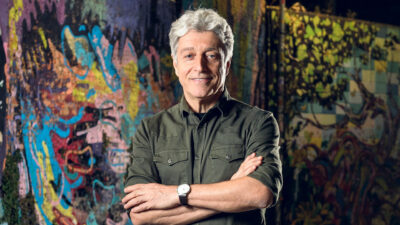 Caco Barcellos com camisa verde escura e braços cruzados em frente a muro com grafite colorido