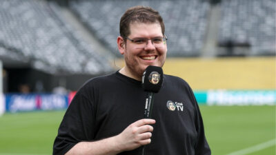 Casimiro Miguel sorrindo, de óculos e camisa preta, segurando microfone da CazéTV em um campo de futebol com arquibancada ao fundo