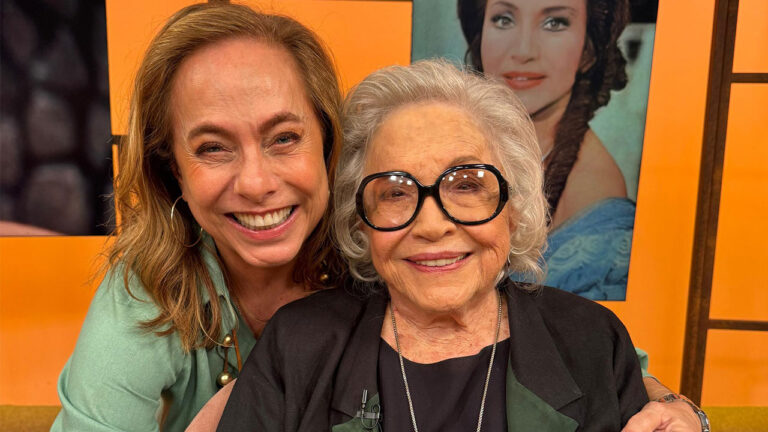 Cissa Guimarães e Nathalia Timberg sorrindo juntas no estúdio do programa Sem Censura com fundo laranja e foto antiga de Nathalia ao fundo