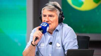 Cléber Machado com fones de ouvido e microfone com espuma azul durante transmissão esportiva da Record
