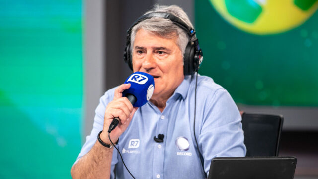 Cléber Machado com fones de ouvido e microfone com espuma azul durante transmissão esportiva da Record
