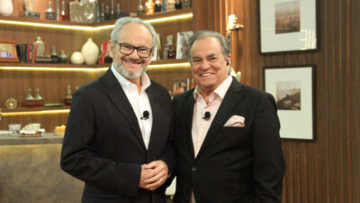 Ernesto Paglia e Ronnie Von sorrindo lado a lado, ambos de terno, em um estúdio com prateleiras ao fundo