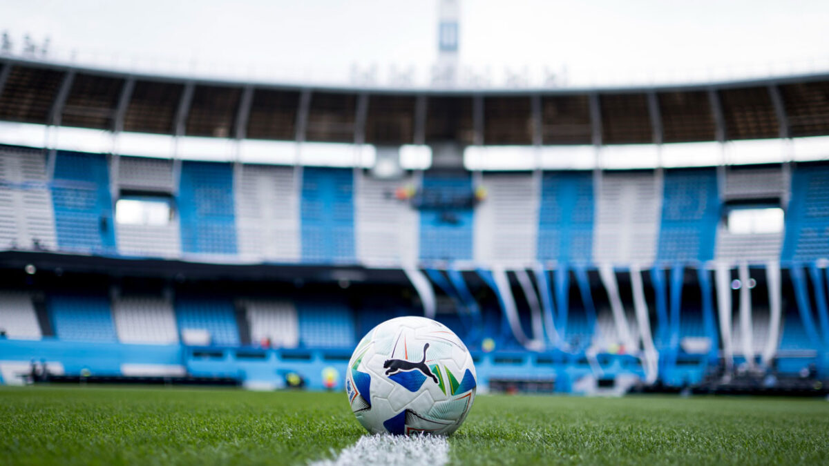 Bola de futebol sobre o gramado na marca central do Estádio Presidente Perón, o El Cilindro, com arquibancadas em azul e branco ao fundo