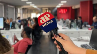 Mão segurando microfone com espuma vermelha e azul exibindo o logotipo da JP News em redação com várias pessoas trabalhando em computadores ao fundo