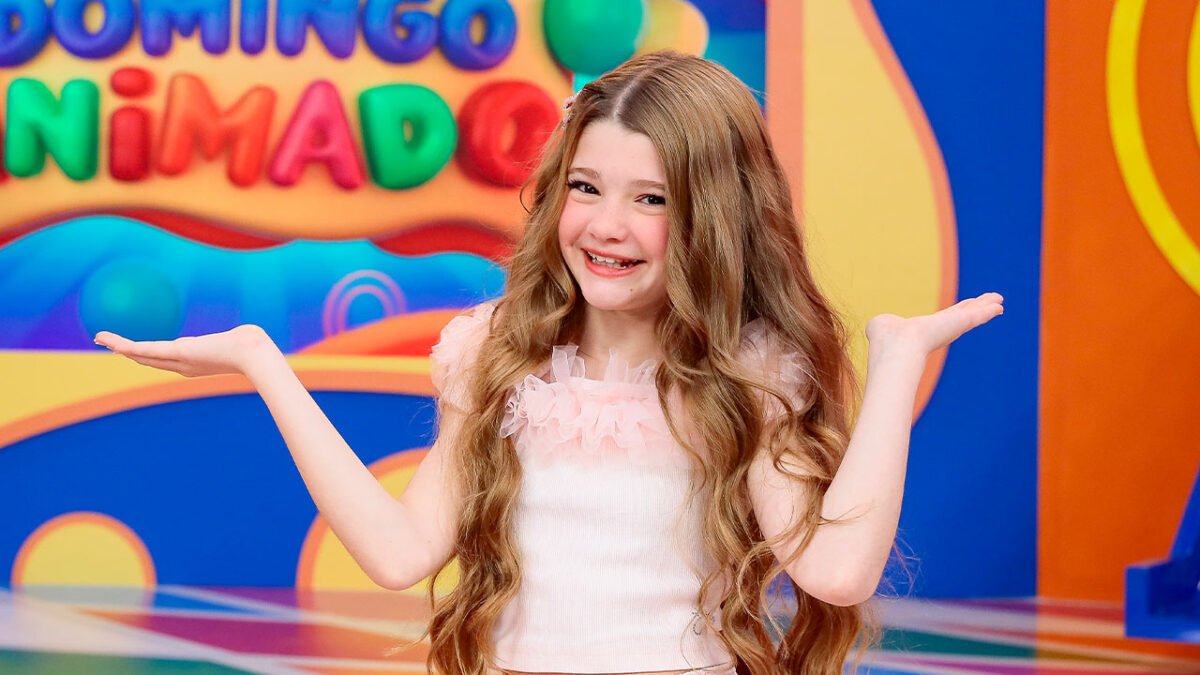A apresentadora Juju Penido, com blusa rosa e cabelo comprido, sorri com as palmas das mãos abertas no estúdio do Domingo Animado