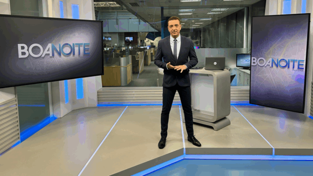 Apresentador Marcelo Rocha de terno escuro em pé no estúdio do Boa Noite Paraná com telões e a redação ao fundo